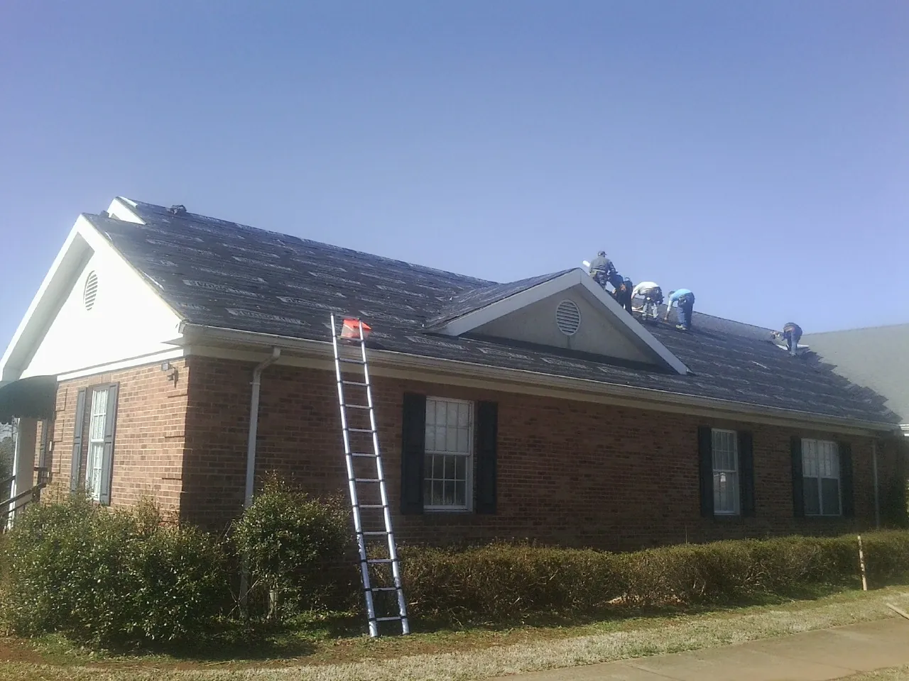 Crew tearing shingles off a brick ranch roof with a ladder set — Emergency Roof Repair work by our crew in Mc Intire