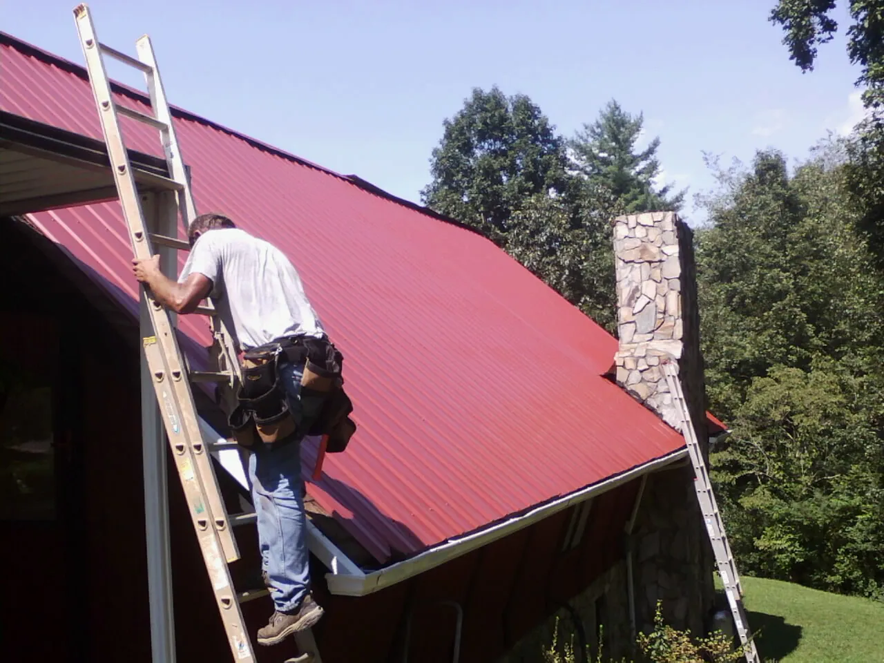 Roofer on a ladder beside a red metal roof and stone chimney — Soffit & Fascia Repair by Roofing Brothers in Mc Intire